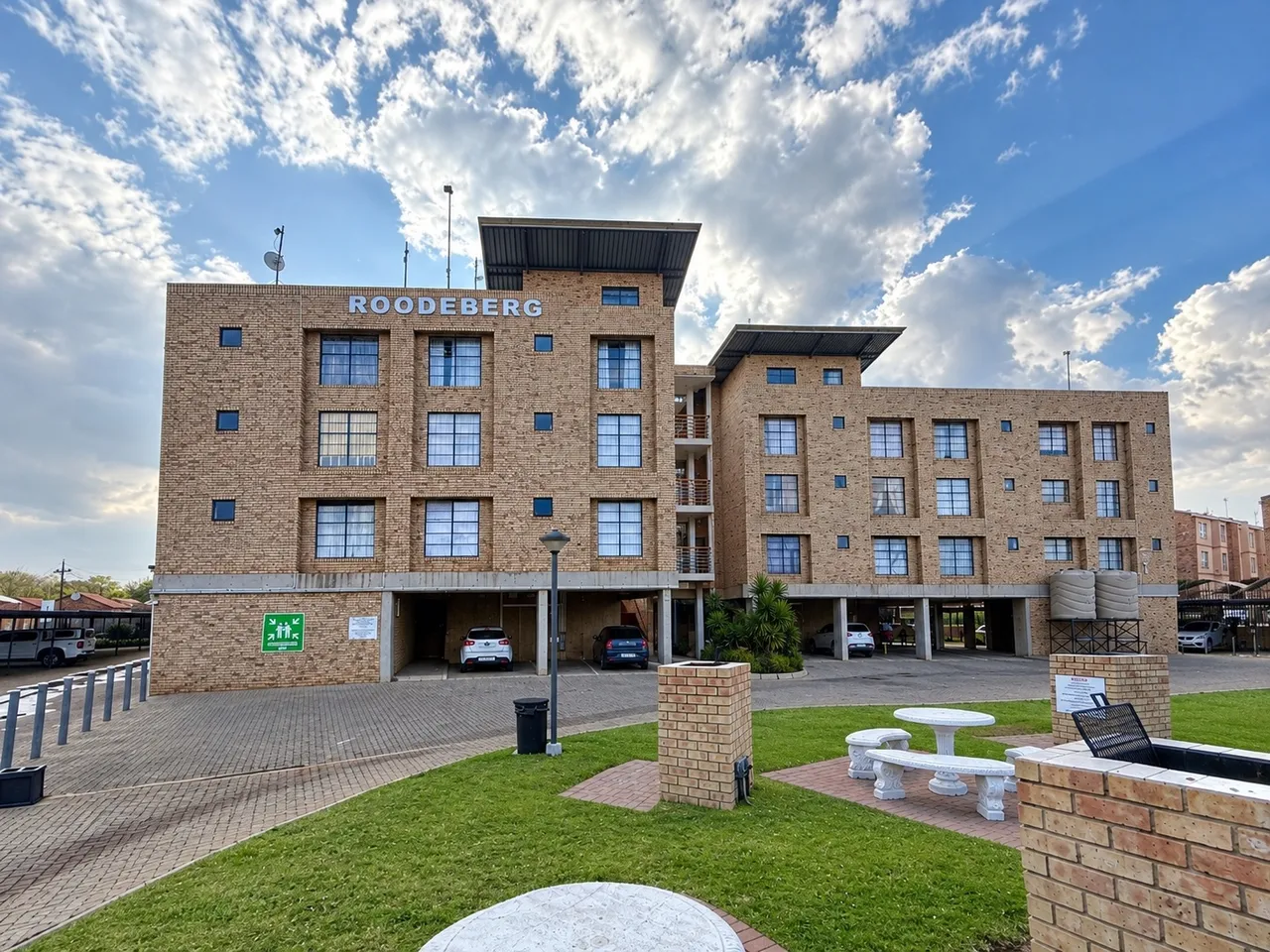 Four-storey face-brick apartment block with the name Roodeberg on its facade, covered parking underneath and a lawn with concrete benches in front, under a blue sky with clouds