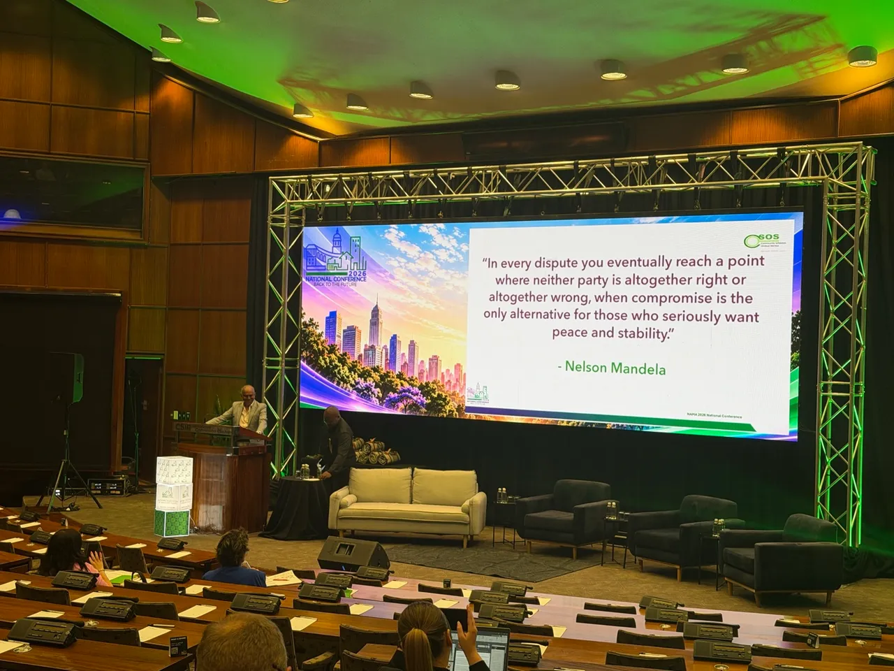 A speaker at a wooden podium in a wood-panelled auditorium beside a large screen showing a CSOS slide with a Nelson Mandela quote on compromise, under the NAMA 2026 National Conference branding, with delegates seated in the foreground