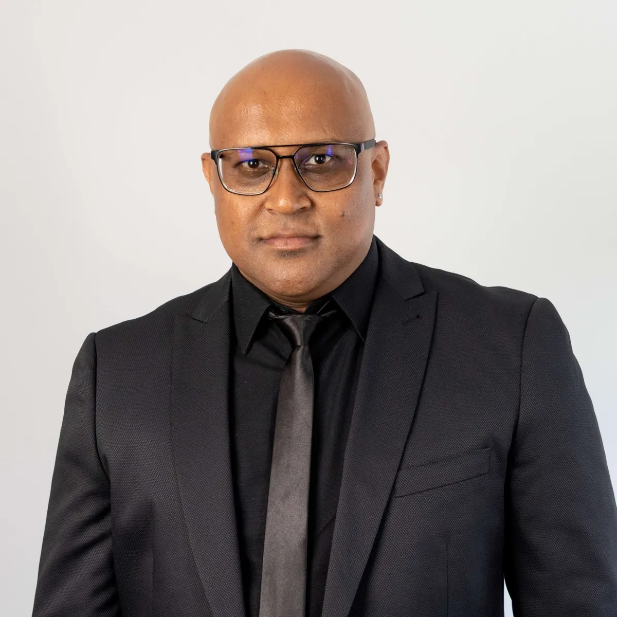 Viresh Persad, Chief Development Officer of Newlyn Group, in a black suit, shirt and tie and dark-framed glasses against a plain light background