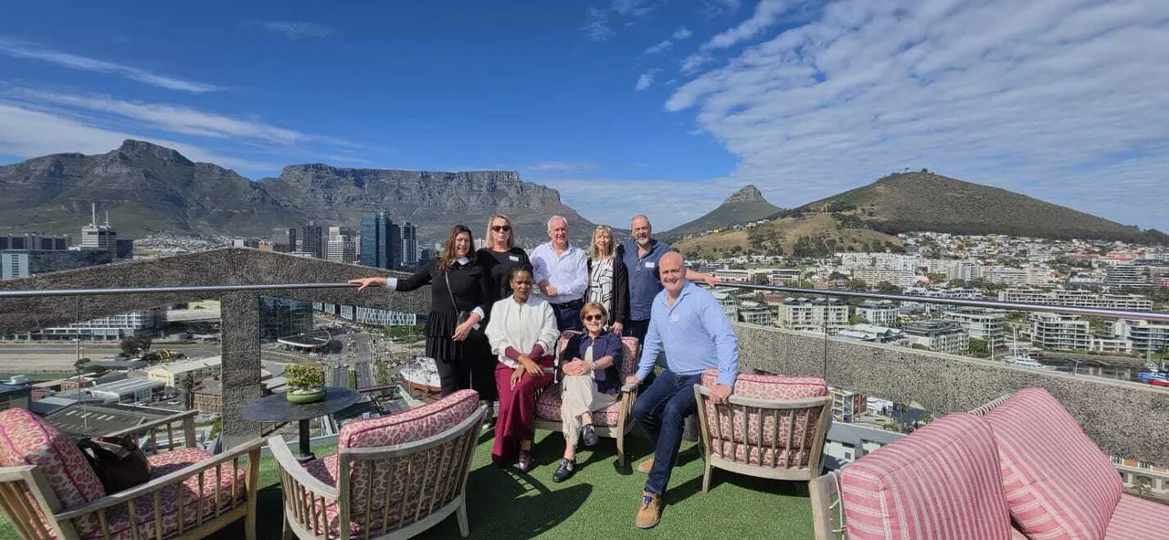 Norbert Sasse, in a white shirt, standing at the back in the middle of a group of eight on a rooftop terrace with pink striped chairs, Table Mountain, Lion's Head and the Cape Town city bowl behind them