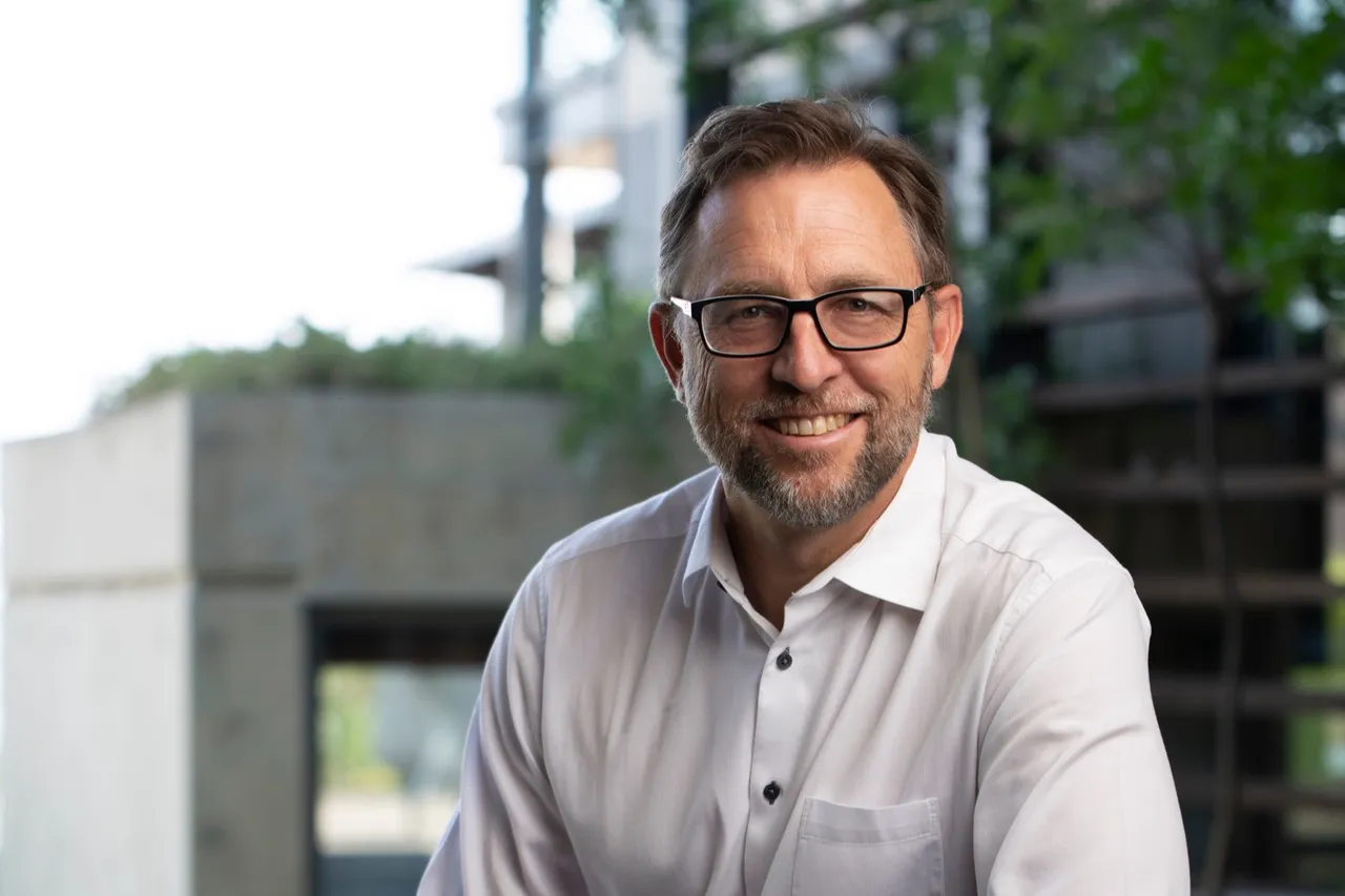 Pieter Lombaard, CEO of the Moolman Group, smiling in glasses and a white shirt with dark buttons, seated outdoors in front of a concrete wall and greenery