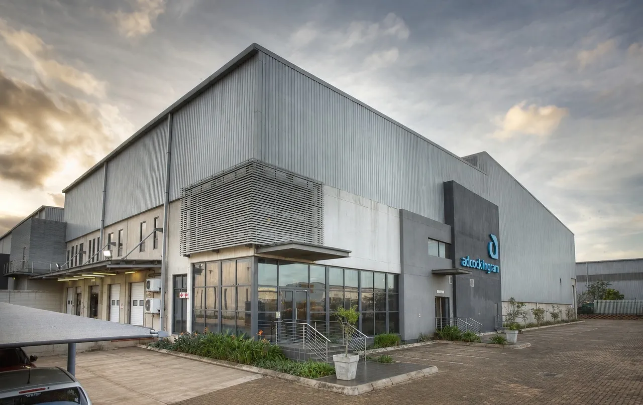 A grey industrial warehouse with a glass-fronted office entrance and Adcock Ingram signage under a cloudy sky