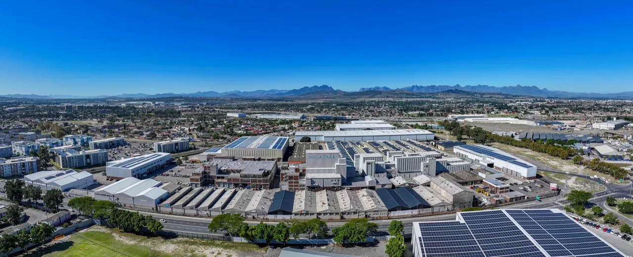 Panoramic aerial view of MegaPark in Bellville: rows of industrial and warehouse buildings, some with solar panels on their roofs, surrounded by apartment blocks under a clear blue sky with mountains on the horizon