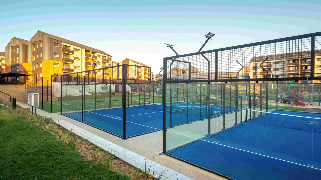Two glass-walled padel courts with blue surfaces at Indawo Lifestyle Apartments, with apartment blocks and a children's playground behind them