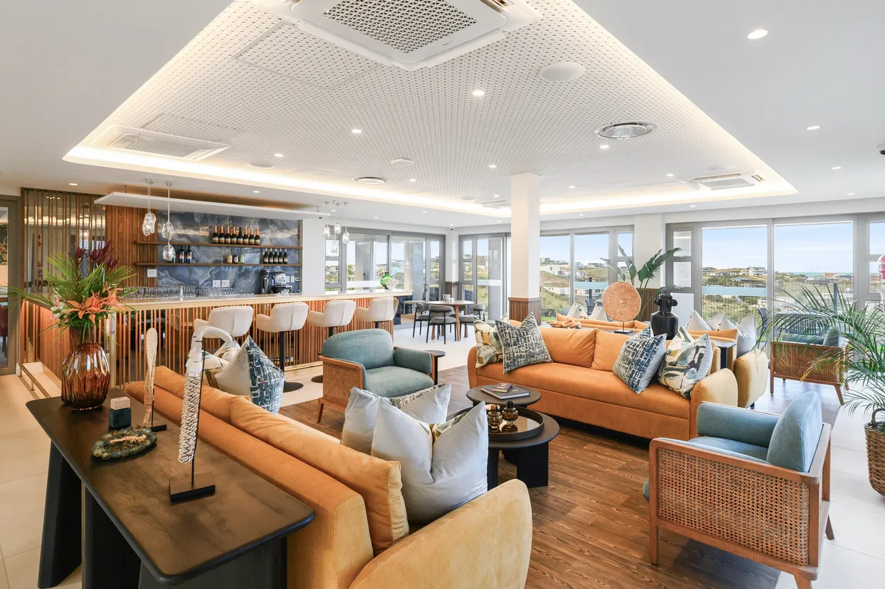 A residents’ lounge and bar with sofas, rattan armchairs and floor-to-ceiling windows looking out over the coastal estate