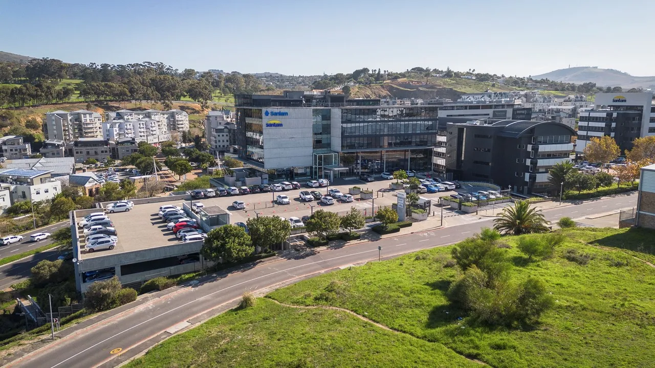 Aerial view of the Santam Precinct in Tygervalley: a glass-fronted office block with Santam and Sanlam signs, a rooftop parking deck and a full parking lot, with trees and hills behind it