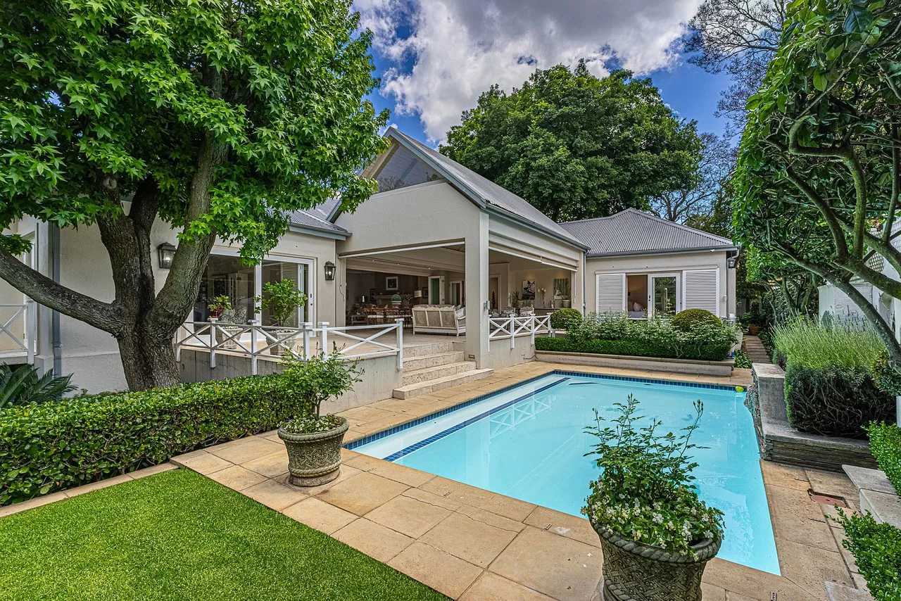 A white single-storey Melrose home with a covered veranda and white balustrades, a rectangular swimming pool edged in sandstone paving, potted shrubs and a lawn, under large trees