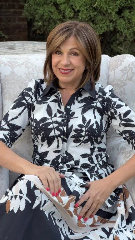 Marina Constas, Director at BBM Attorneys, smiling, with a brown bob and red lipstick, in a black and white leaf-print shirt dress seated on a white patterned sofa