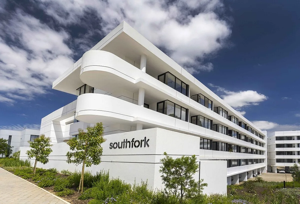 A white, curved four-storey apartment block with Southfork signage on a garden wall, under a blue sky with clouds