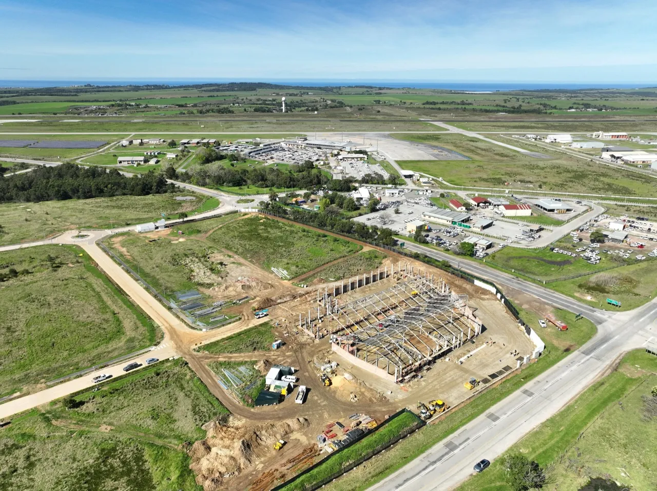 Aerial view of the GTX Industrial Park building site next to George Airport: the steel frame of a large industrial building going up on cleared land, with the airport terminal, runway and the sea in the distance
