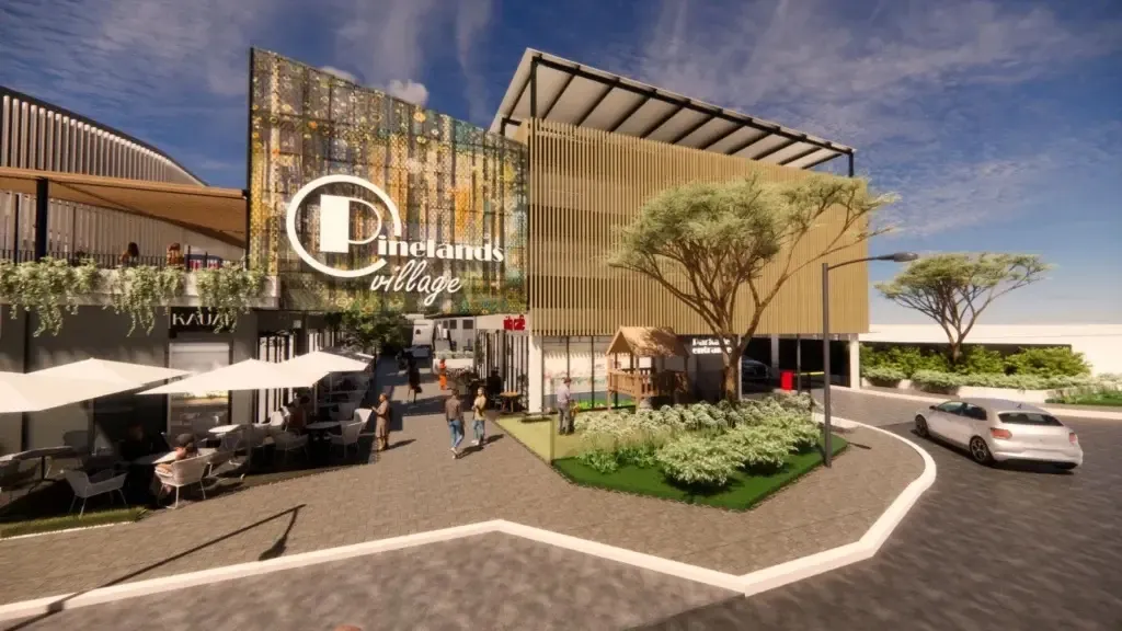 Architectural render of Pinelands Village: the Pinelands Village sign on a patterned facade above a paved walkway with restaurant tables and umbrellas, a wooden children's play structure on a lawn and a car passing