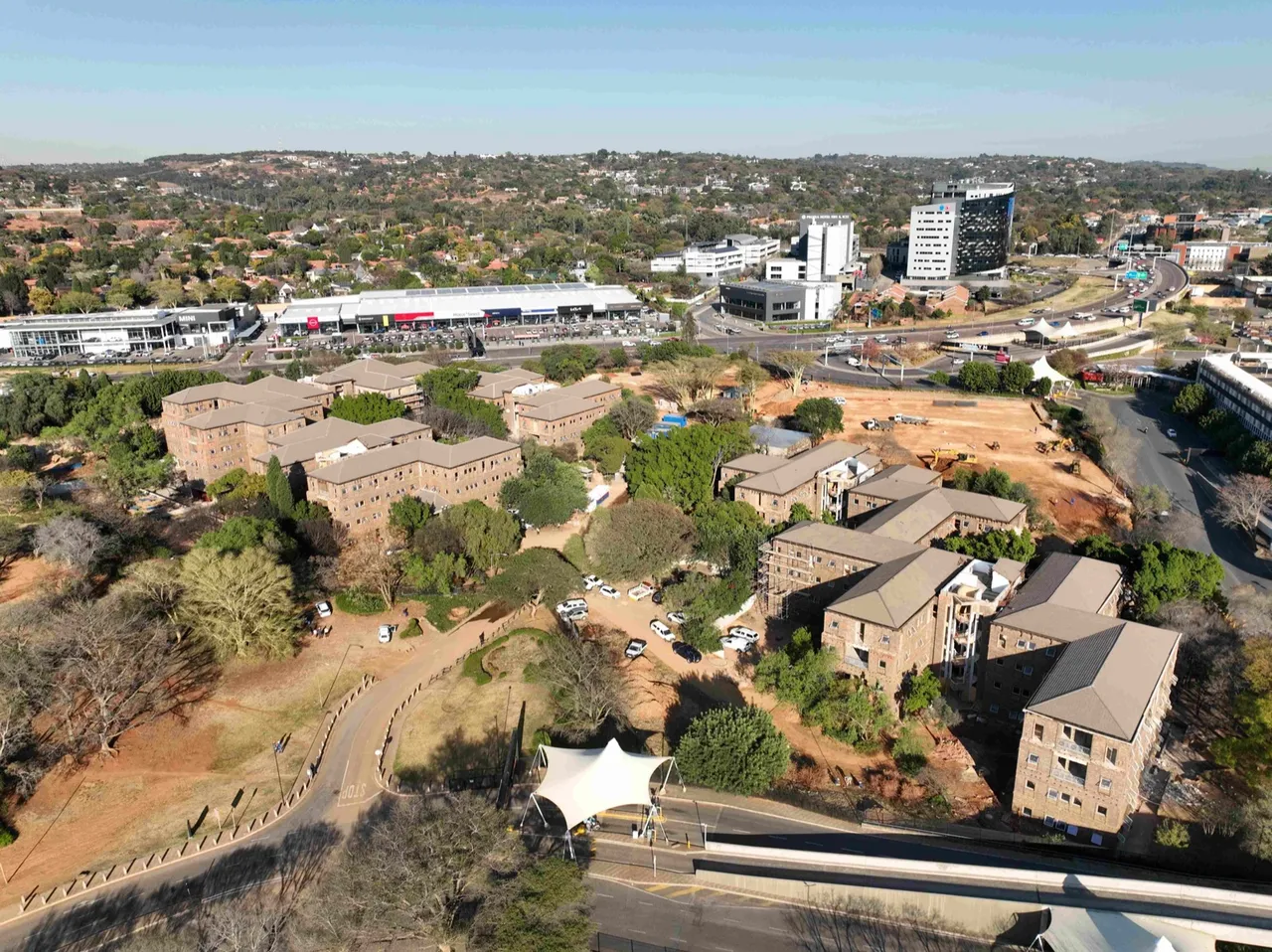 Aerial view of the former Menlyn office park: the existing face-brick office blocks among mature trees, with a cleared construction site beside them and Menlyn's shops and office towers behind