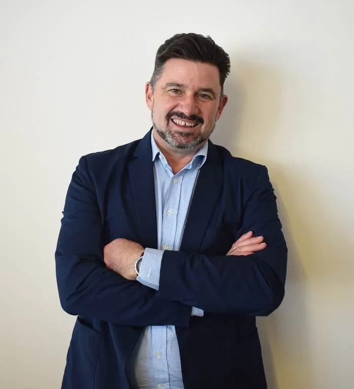 Rhys Dyer, CEO of the ooba Group, smiling with his arms folded, in a navy blazer over a light blue checked shirt against a plain white wall