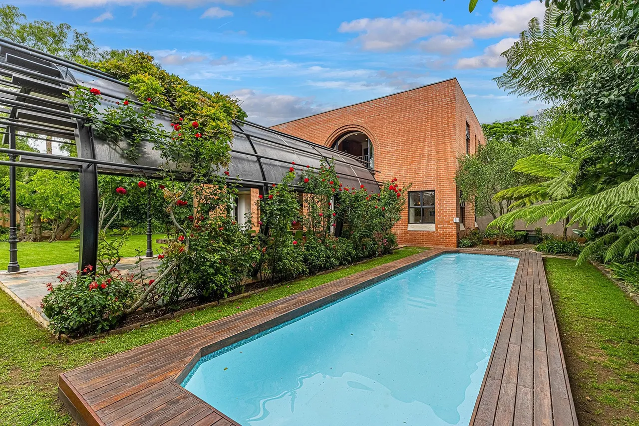 A red-brick Westcliff home with an arched window, a long swimming pool with timber decking, and a steel-and-glass pergola covered in red roses beside a lawn