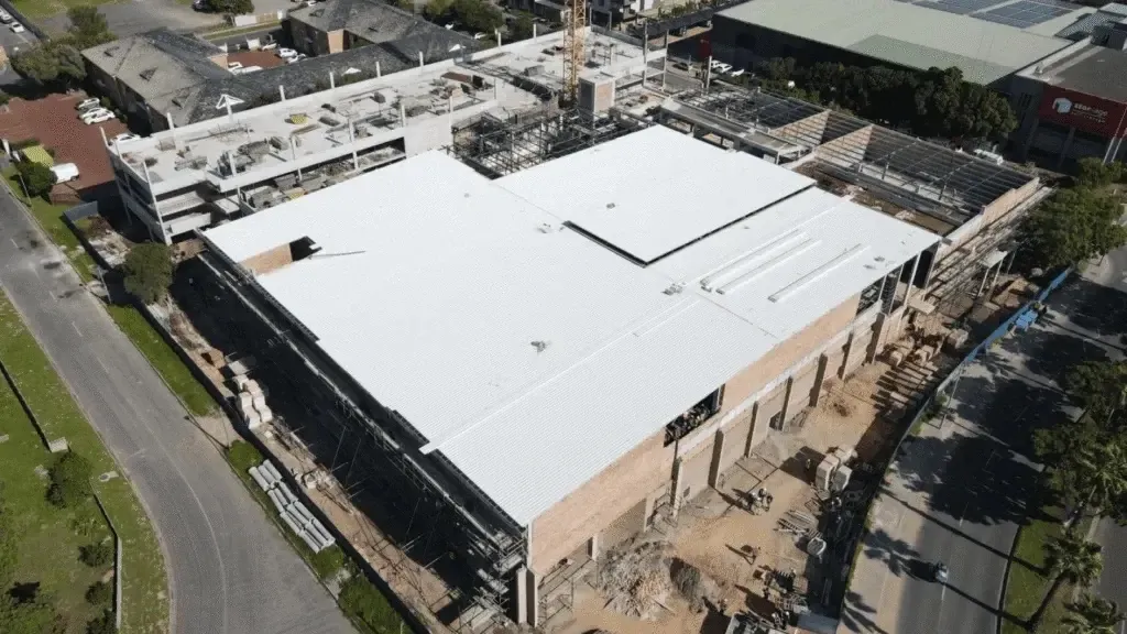 Aerial view of the Pinelands Village construction site: a large white-roofed brick building with scaffolding and building materials along its sides, a crane and an unfinished parking deck behind, between tree-lined roads