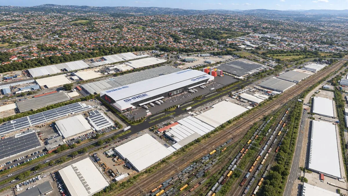 Artist's impression from the air of a large Shree-branded logistics warehouse among industrial buildings and solar-panelled roofs, beside a railway yard, with suburbs and hills behind