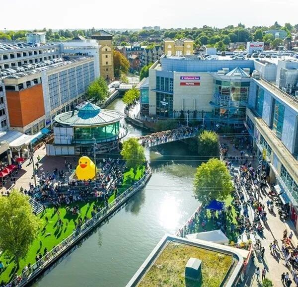 Aerial view of a busy shopping destination built around a canal, with crowds on a green lawn beside a giant yellow duck and glass-fronted retail buildings