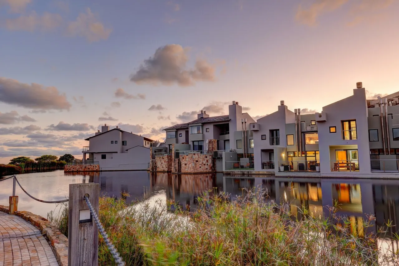 Grey-and-white apartments with lit windows reflected in a still lake at dusk, with reeds and a rope-railed walkway in the foreground