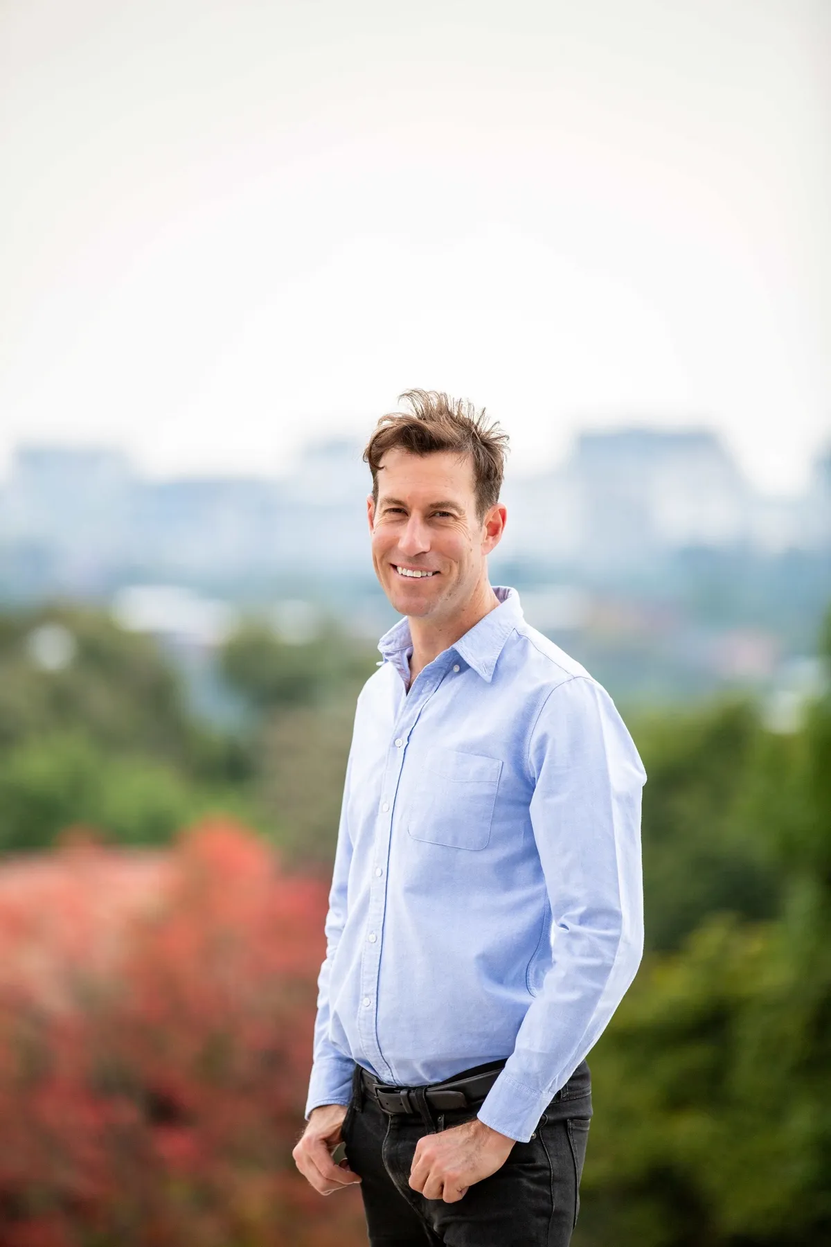 Justin Blend, Director of Africrest, smiling in a light blue shirt and dark jeans, standing outdoors with trees and a blurred city skyline behind him