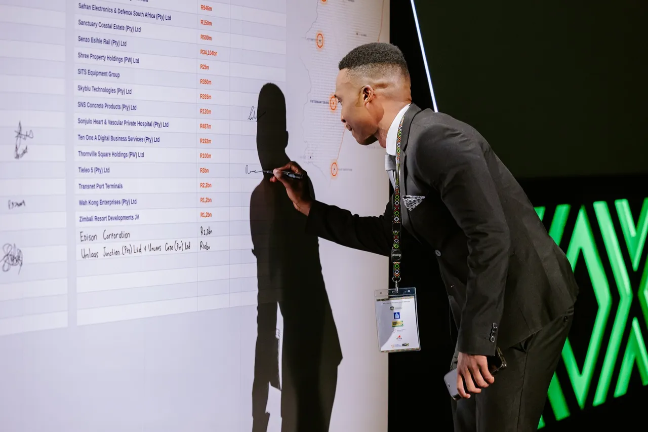 A man in a dark suit and conference lanyard signs the investment pledge wall beside the line "Tieleo 5 (Pty) Ltd, R3bn", with a map of KwaZulu-Natal on the screen behind the list