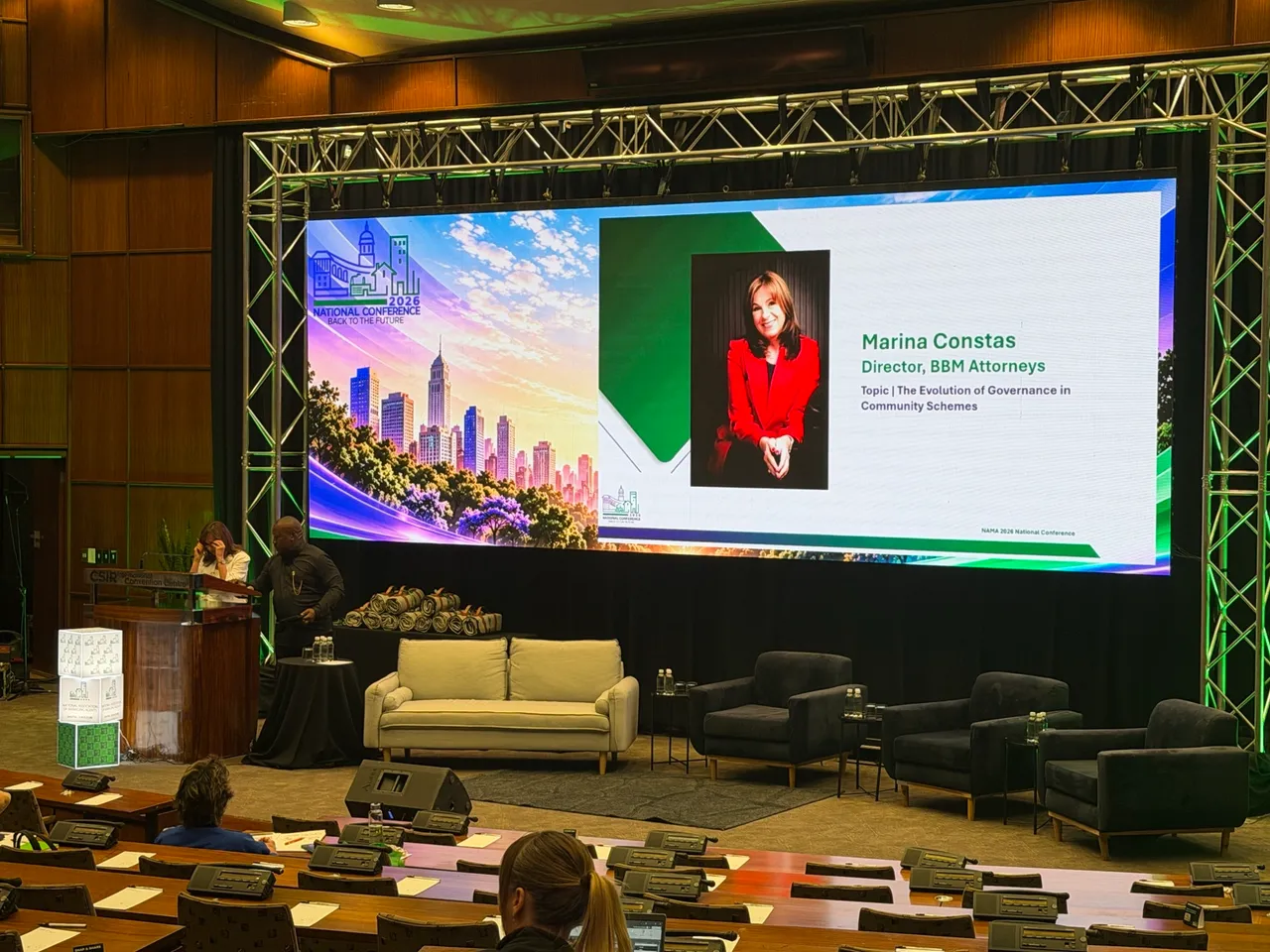 The stage at the NAMA 2026 National Conference at the CSIR International Convention Centre, with a large screen showing a slide for Marina Constas, Director, BBM Attorneys, and a sofa and armchairs on stage