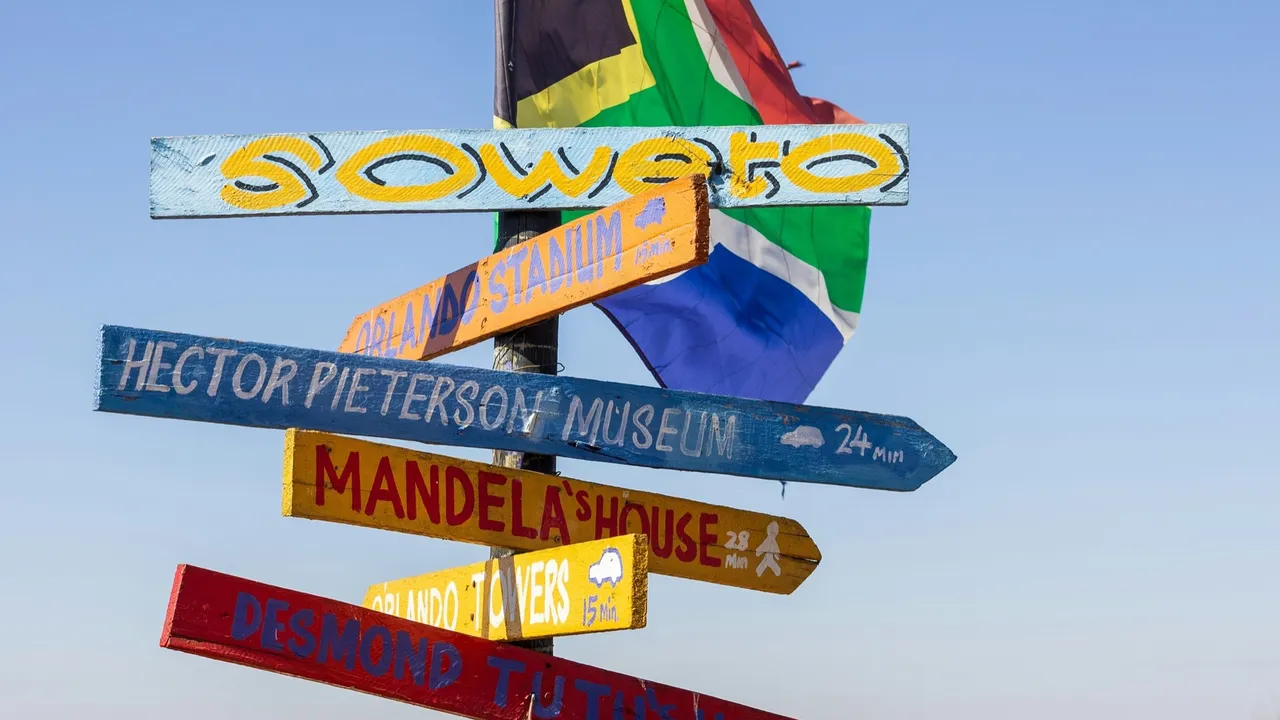 A colourful hand-painted signpost reading Soweto, with arrows to Hector Pieterson Museum, Mandela's House, Orlando Towers, Orlando Stadium and Desmond Tutu's house, in front of a South African flag and a blue sky