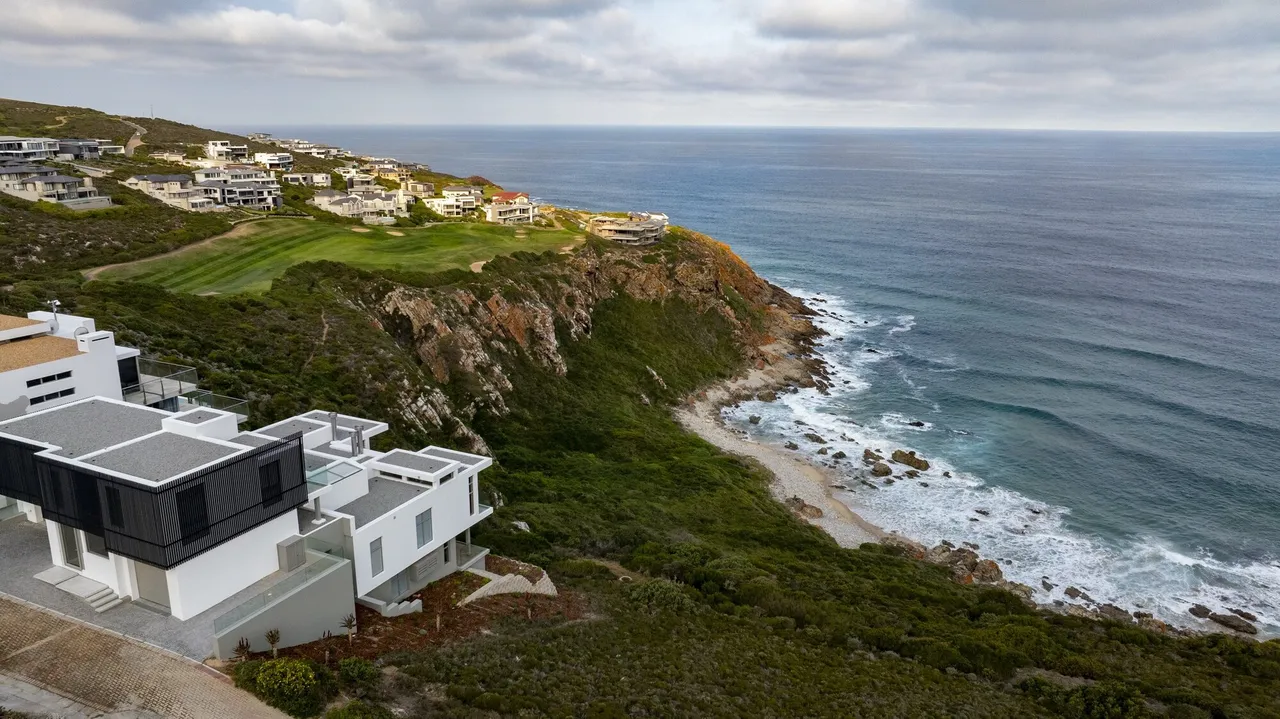 Aerial view of a new white clifftop home at Pinnacle Point, with a golf fairway, more homes on the headland and the ocean beyond