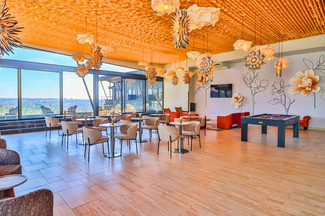 The Maestro Sky Lounge at an Africrest development: a residents' lounge with café tables, orange sofas and a pool table, paper flower lights under a wooden slatted ceiling and floor-to-ceiling windows overlooking the city