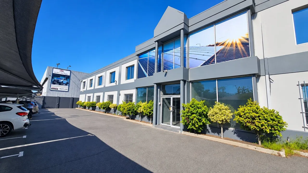 Two-storey grey-and-white industrial building with tall windows showing a solar-panel graphic, a row of shrubs along the front and a shaded parking area, in Airport Industria, Cape Town