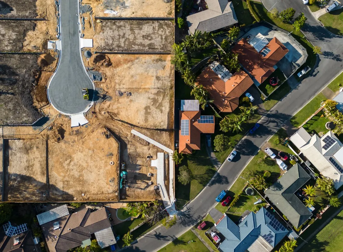 Aerial view of a new housing development, with cleared plots and a freshly tarred cul-de-sac beside an established suburb of tiled-roof houses with solar panels