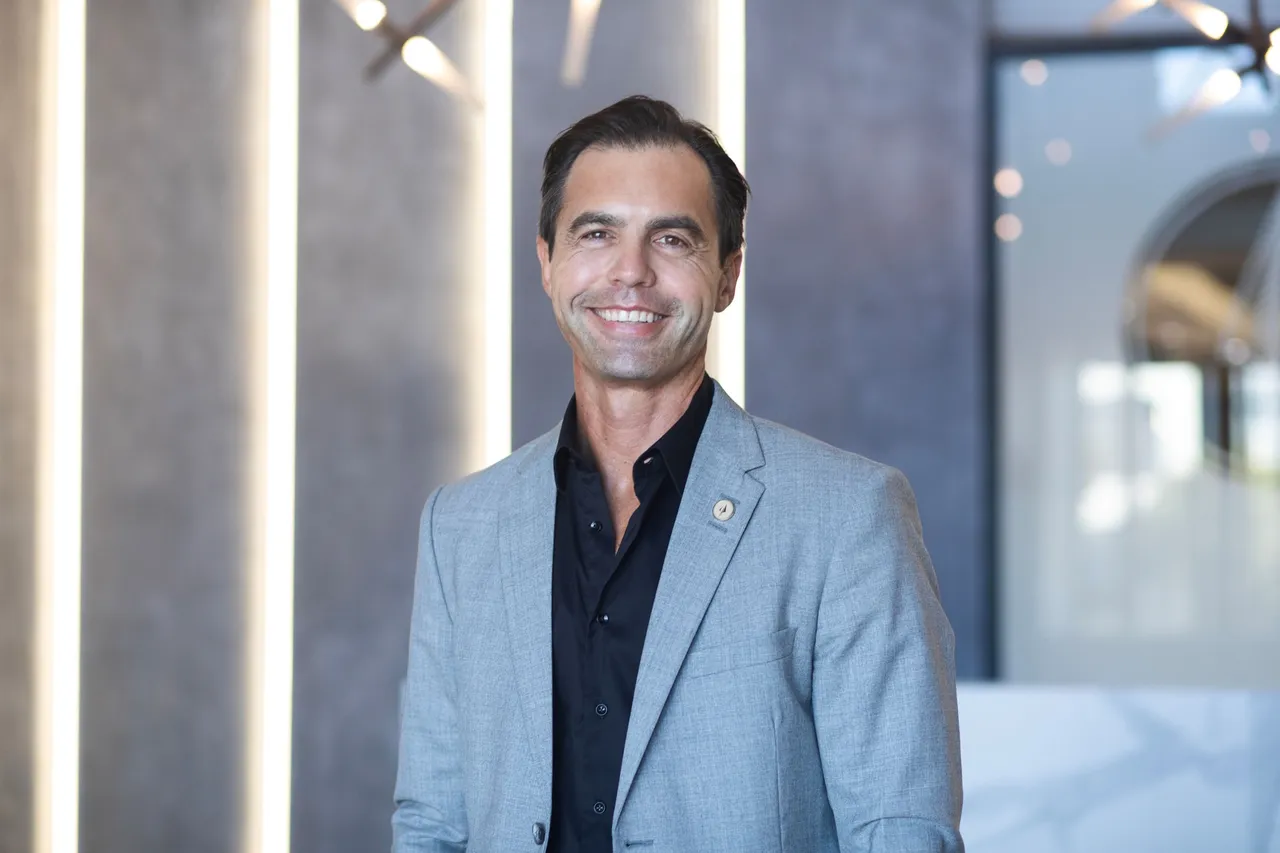 Quintin Rossi, CEO of Spear REIT, smiling in a light grey blazer and black open-collared shirt in a modern lobby with vertical light panels behind him