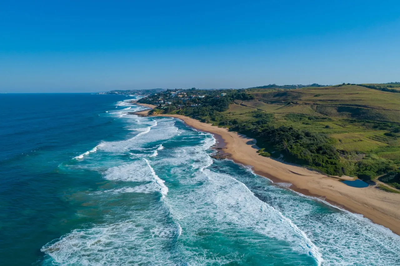 Aerial view of the KwaZulu-Natal North Coast: surf rolling onto a long sandy beach below green hills and coastal homes